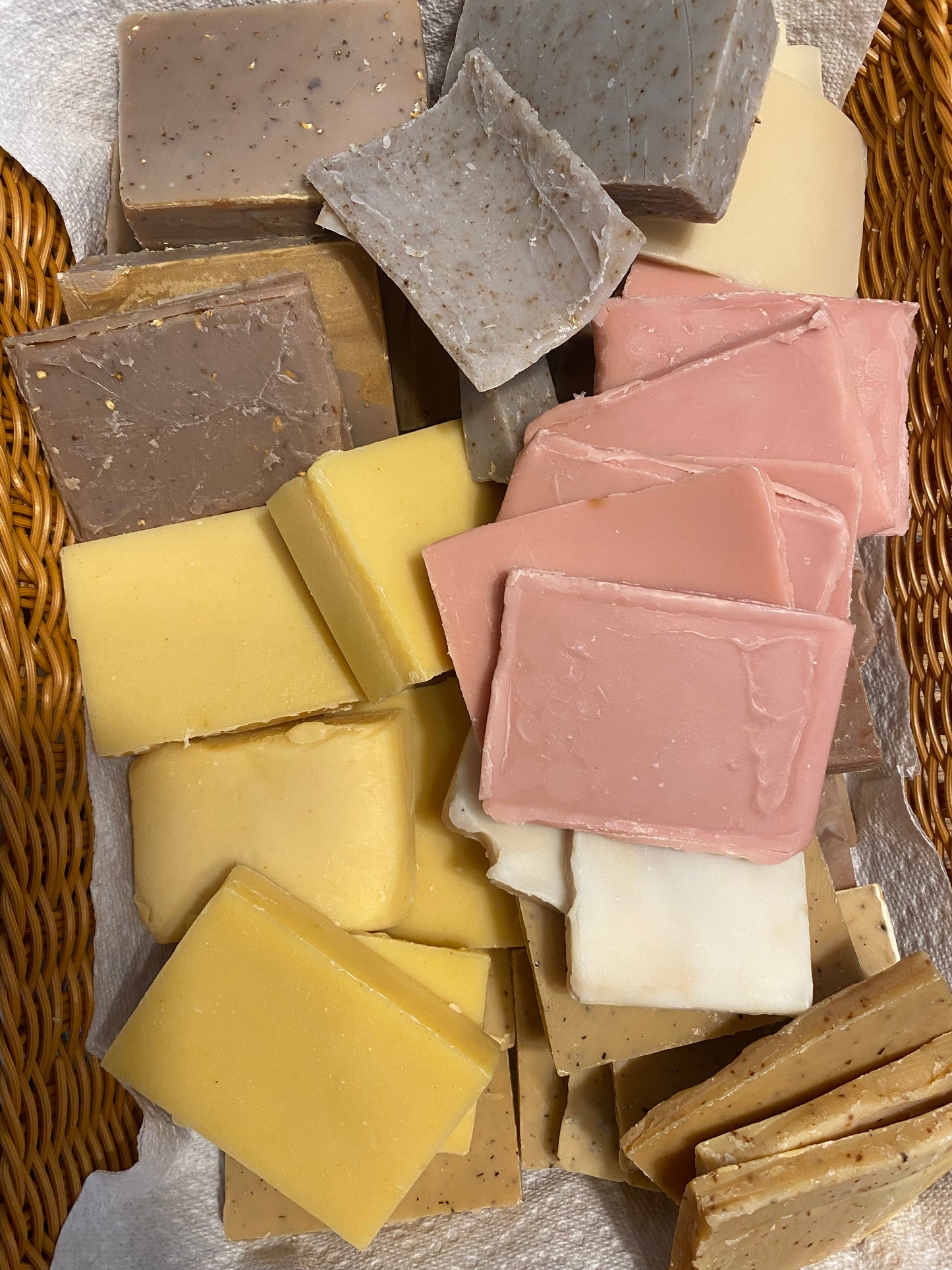 An assortment of handmade rectangular bars of soap in various colors including yellow, pink, brown, and gray, arranged in a wicker basket on a white cloth.