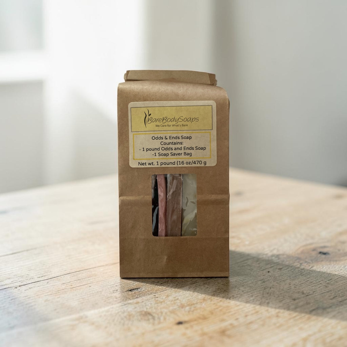 A brown kraft paper bag of  Odds and Ends of soap, standing on a light wooden table with a soft-focus indoor background.